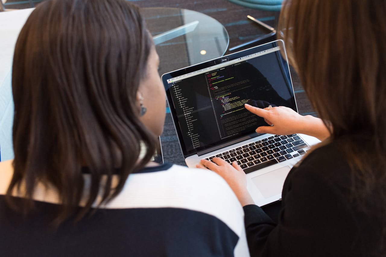 heros-img Two women working together on software programming indoors, focusing on code.